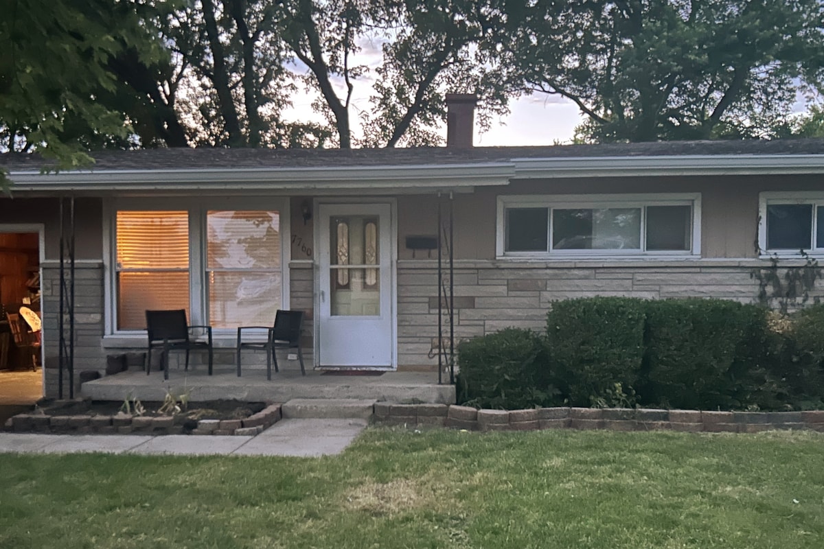 A single-story home is surrounded by green landscaping, featuring a stone exterior and well-maintained bushes. Two chairs are positioned on the porch, ideal for relaxation. Windows allow natural light to flow into the space, contributing to a welcoming atmosphere.