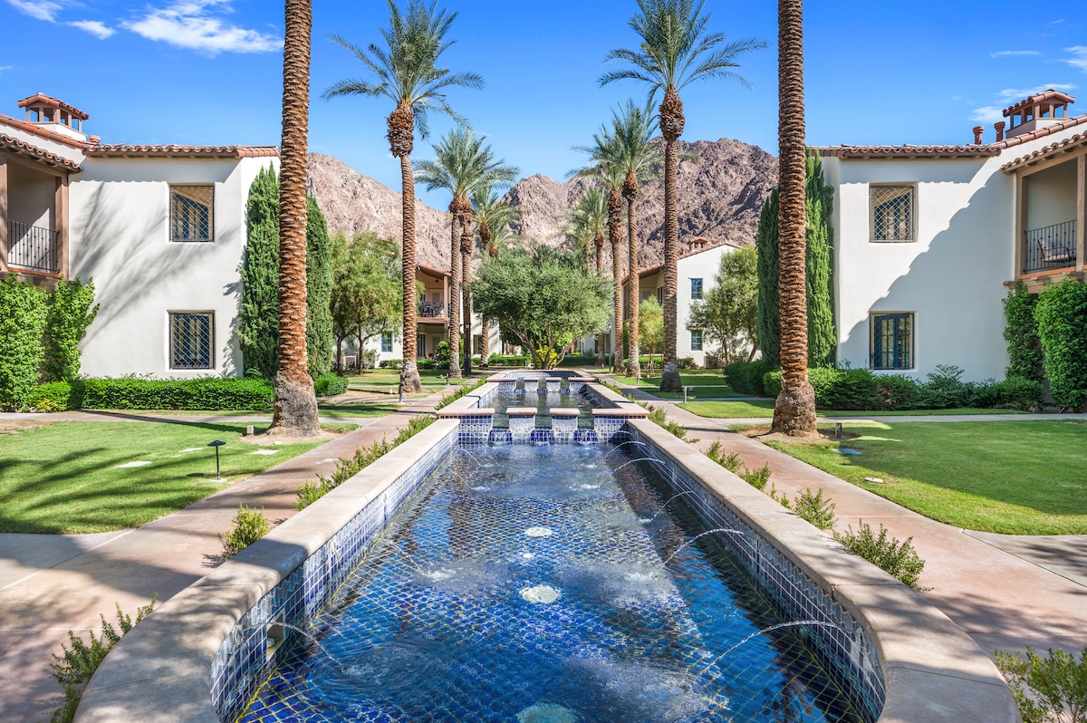 The image captures a serene landscape featuring a tranquil water feature lined with tiles, flowing through a well-maintained garden. Palm trees are positioned along the pathway, with elegant white buildings visible in the background, framed by lush greenery and mountains in the distance.