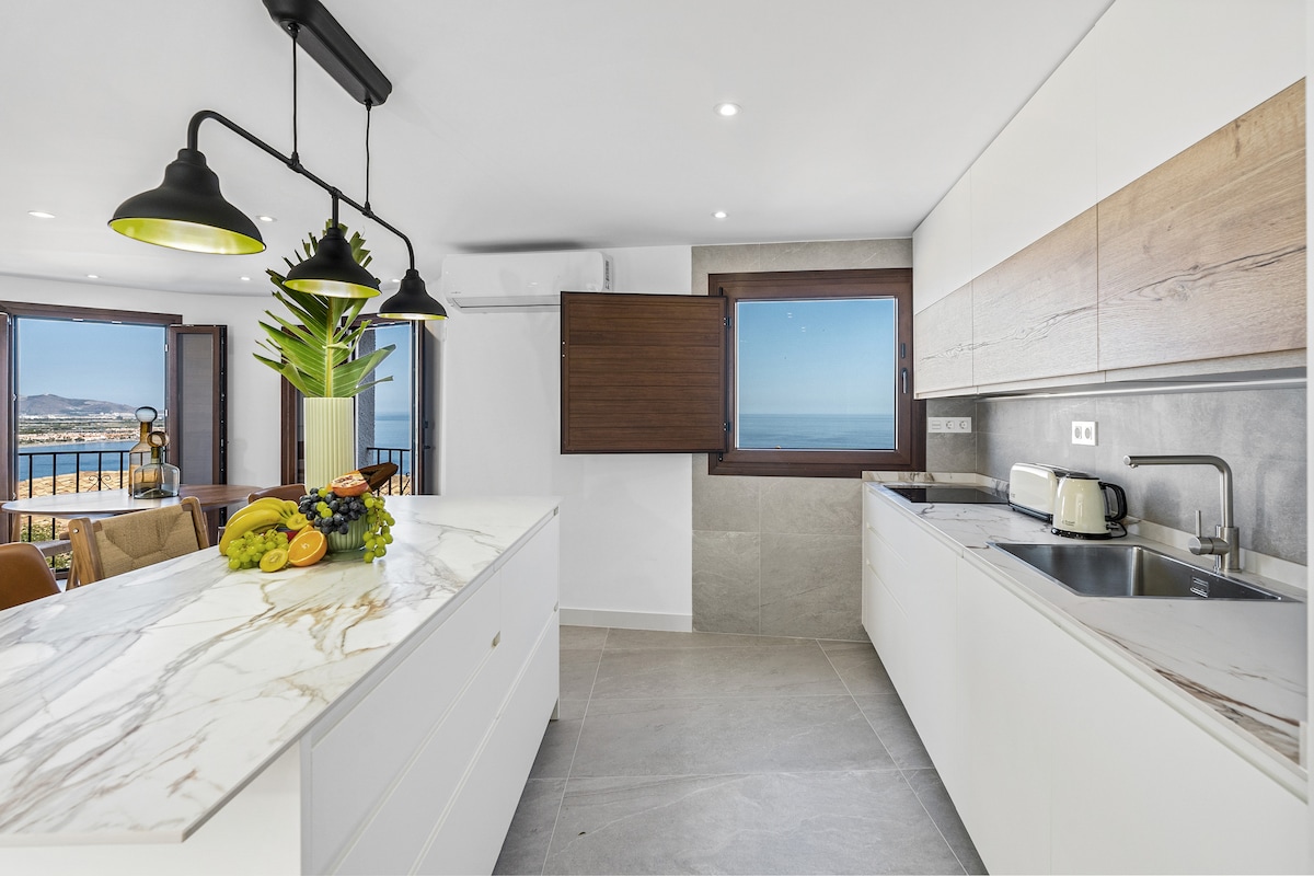 The modern kitchen features sleek white cabinetry and a spacious marble countertop. A selection of fresh fruits is arranged on the counter. Expansive windows offer panoramic views of the sea, while stylish pendant lights add illumination to the space.