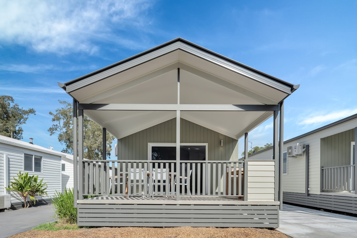 The cabin exterior features a symmetrical pitched roof and a spacious front verandah, adorned with outdoor seating. Lush greenery is visible in the surrounding landscape, while the front facade showcases a mix of light and dark siding. Ample natural light filters through the main entrance.