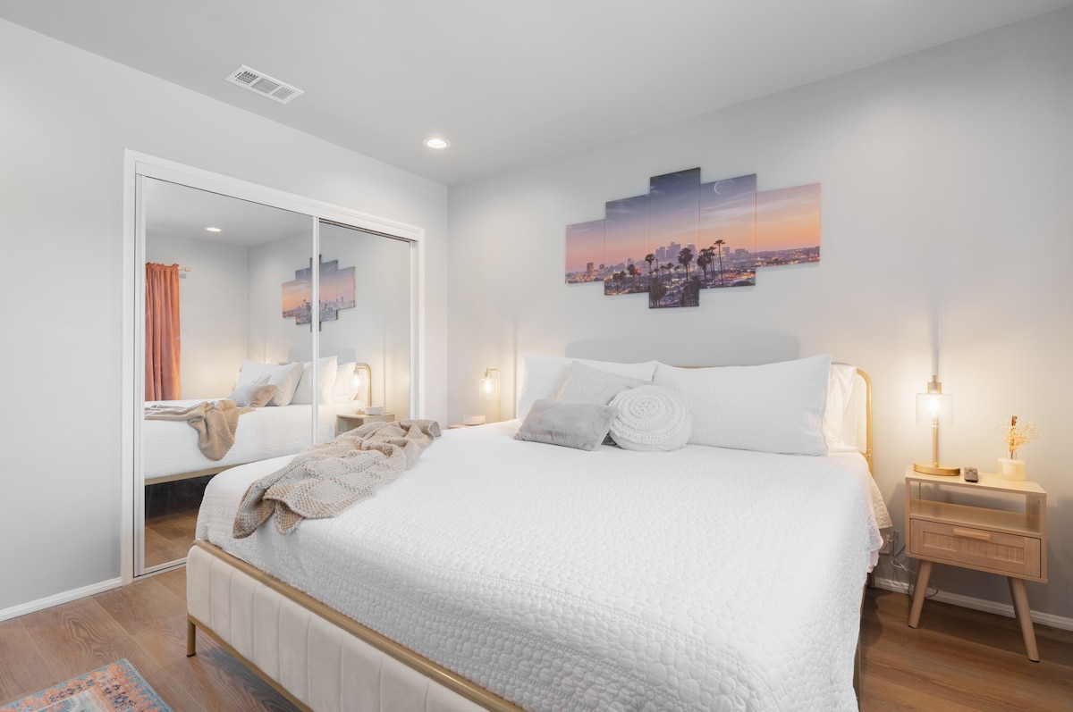 A modern bedroom features a queen-sized bed dressed in white bedding, accompanied by decorative pillows. A large mirror reflects the inviting space. Soft lighting from bedside lamps adds warmth, while a scenic artwork of a city skyline hangs above the bed. Light wood flooring complements the room.