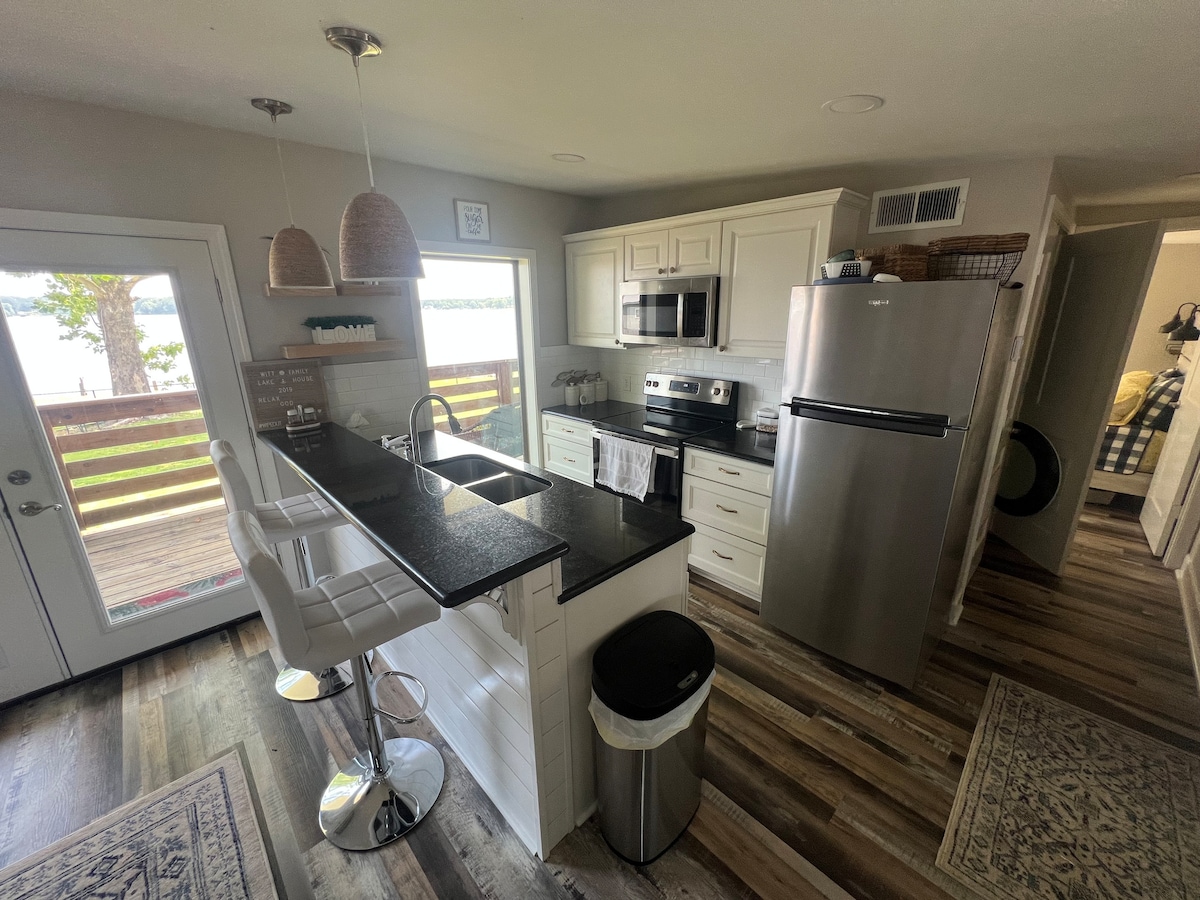 A modern kitchen is visible, featuring white cabinetry and a dark granite countertop. Bar stools are positioned at the island. A stainless steel refrigerator and appliances are included. Natural light enters through a sliding glass door, revealing a view of the water outside.