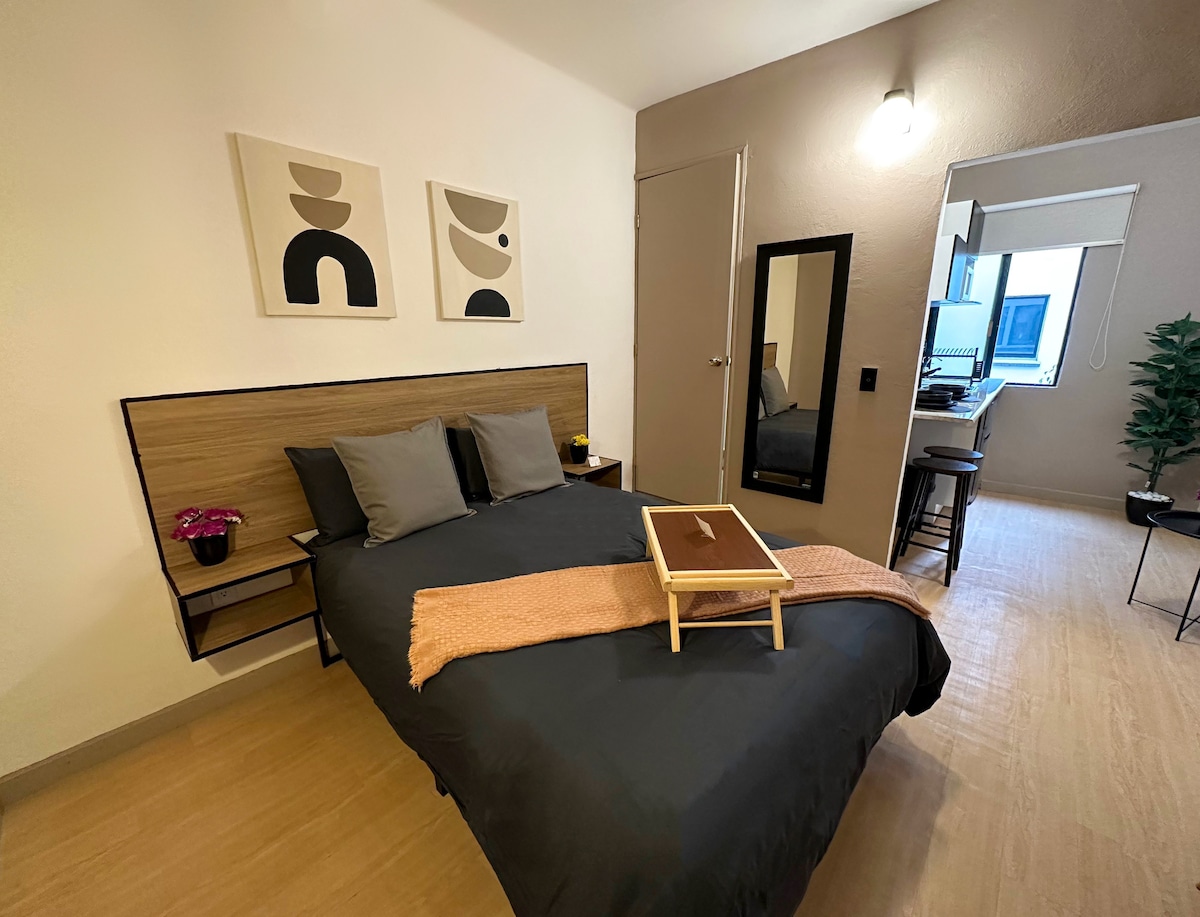 A spacious sleeping area is presented with a dark bedding set on a generous bed, complemented by two neutral-toned artworks mounted on the wall. A soft throw is casually arranged on the bed, and a small wooden table is positioned at the foot of the bed.