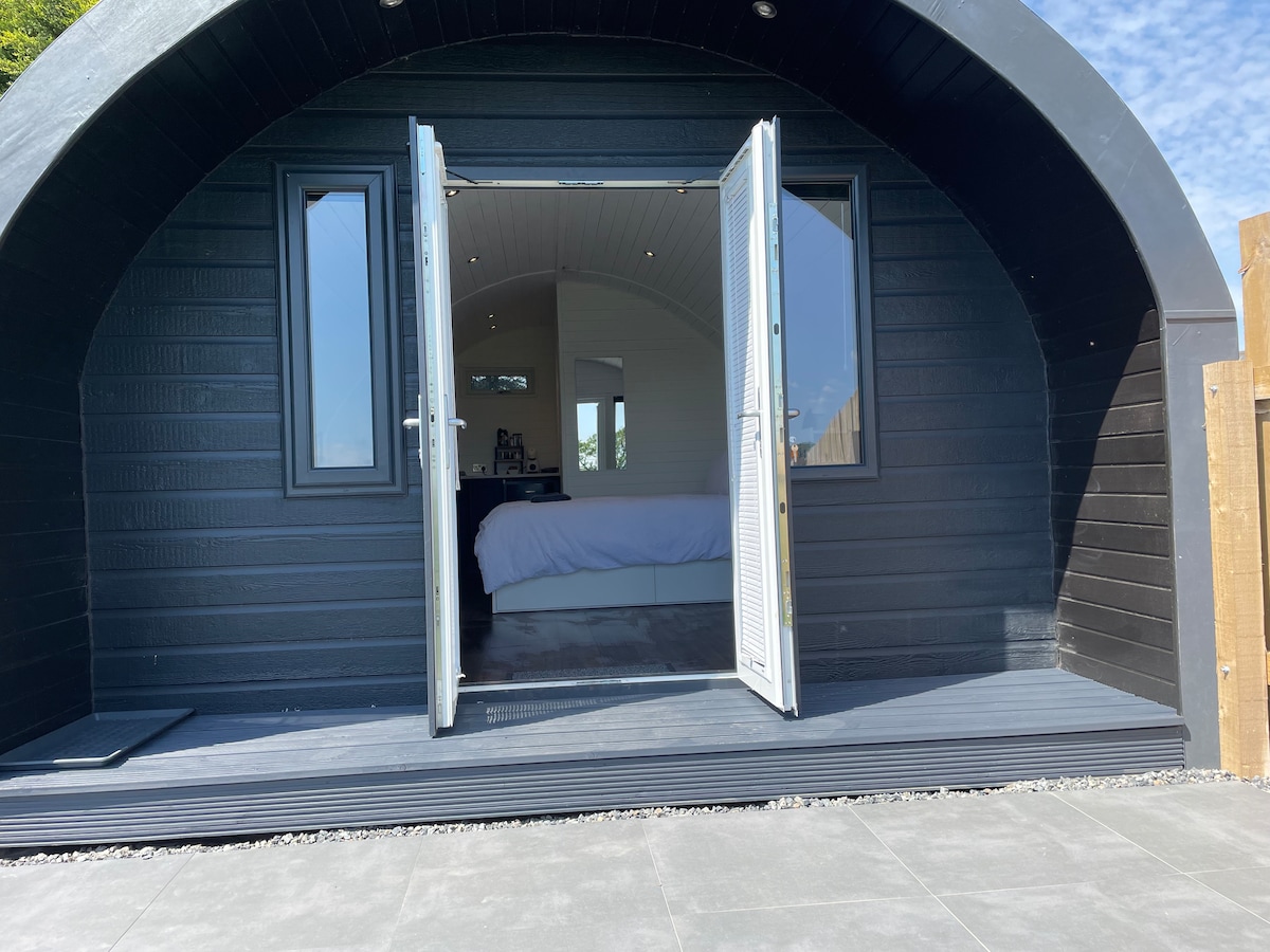 A modern pod is showcased with open double doors revealing a light interior. A spacious bed is visible inside, while a covered wooden deck extends outside. The exterior features a sleek black design with large windows allowing for natural light.
