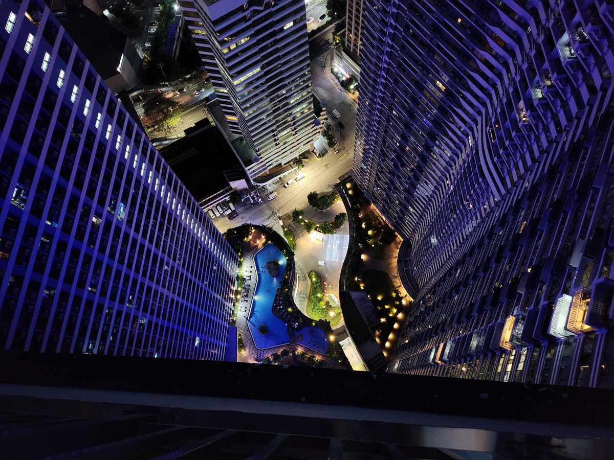 An aerial view of the vibrant Makati CBD at night, showcasing the illuminated resort-style swimming pool and landscaped gardens below. Surrounding skyscrapers are bathed in blue hues, while streets glisten under the city lights, creating a lively urban atmosphere.