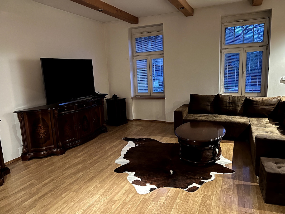 A living room is presented featuring a large flat-screen TV mounted on a dark wooden cabinet. A spacious brown sectional sofa is positioned around a circular coffee table. Natural light filters through two windows, reflecting off the polished wooden flooring, complemented by a dark rug.