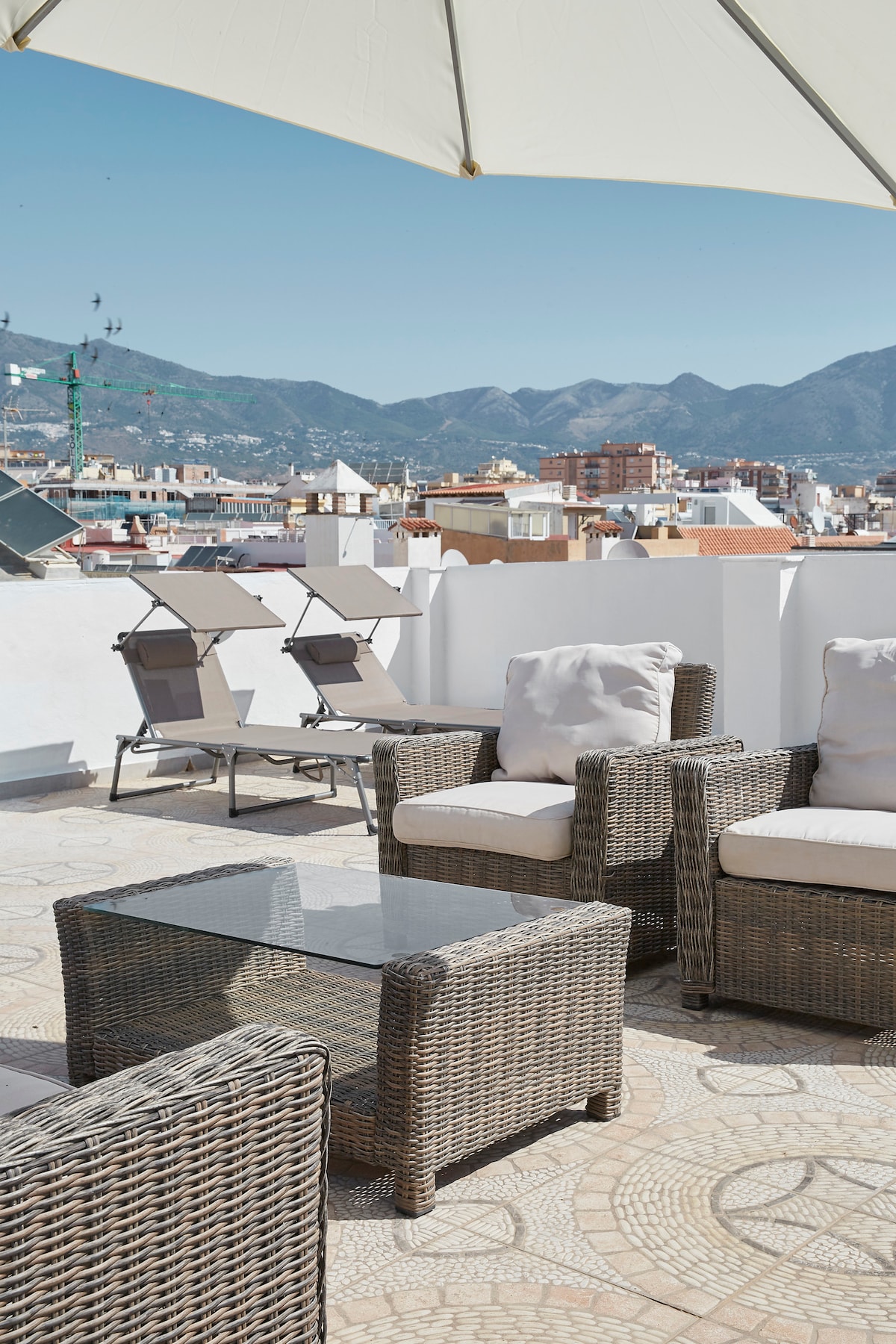 A spacious rooftop terrace is shown, featuring a set of wicker armchairs with soft cushions surrounding a glass-top table. Two lounge chairs are positioned for sunbathing, with a clear view of the city skyline and mountains in the distance, under a bright blue sky.