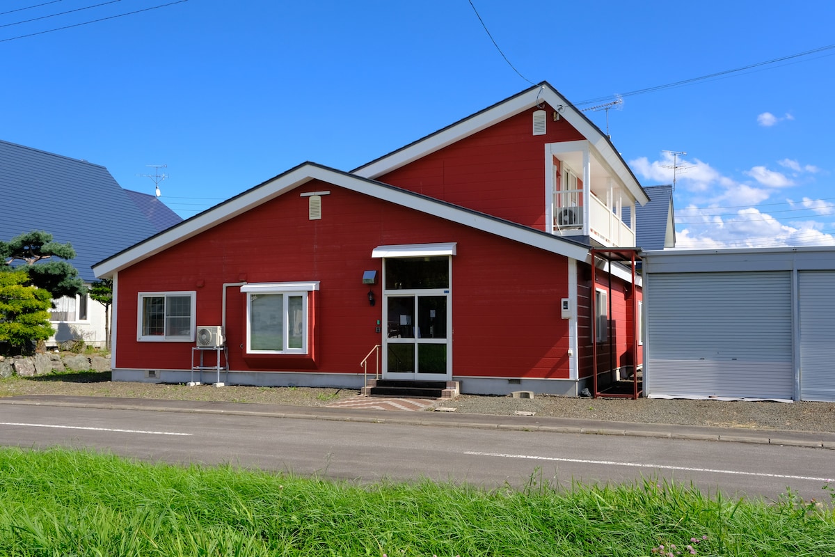 A red, two-story house is presented with white accents and a welcoming entrance. Large windows allow natural light to enter. Grass and a road are visible in front, showcasing the property's accessibility. The exterior features a well-maintained driveway and adjacent structures.