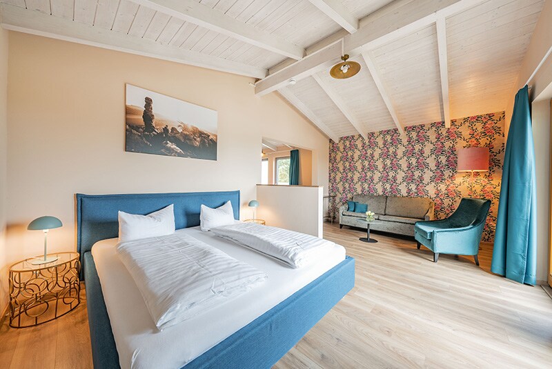 A bright room features a spacious bed with white linens against a backdrop of floral wallpaper. A seating area is provided with an armchair and a sofa, complemented by a soft blue color scheme. Natural light enters through large windows, highlighting the wood ceiling.