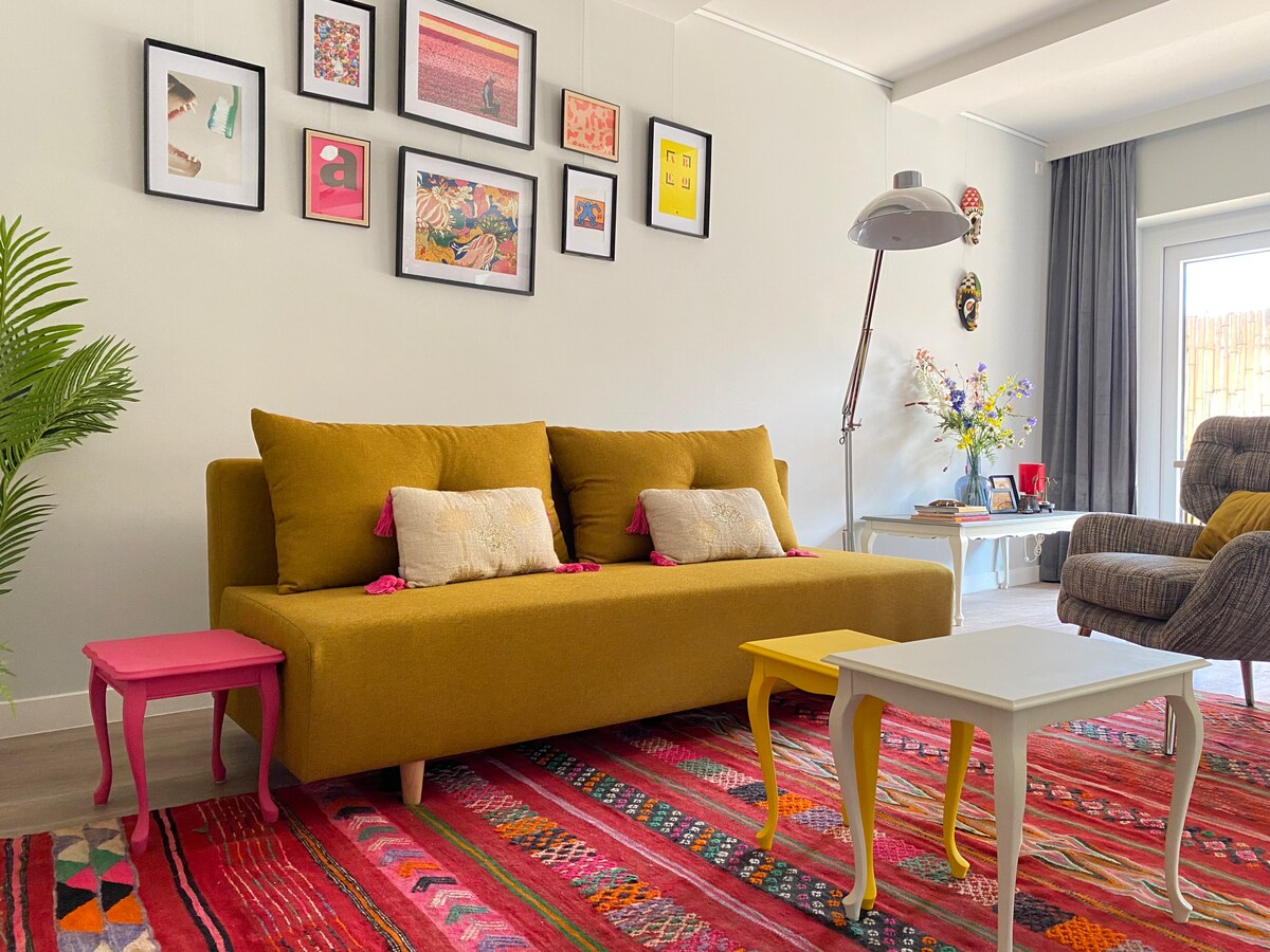 A vibrant living area is highlighted by a yellow sofa adorned with decorative pillows. A colorful area rug features intricate patterns beneath the seating. Small accent tables in pink and yellow complement the stylish decor. A gallery wall with framed art pieces adds visual interest.
