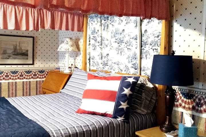 Habitación de abajo (sin subir escaleras) con una cama queen y un sofá cama, un tema rojo blanco-azul. Una ventana de la bahía da al oeste, con vista al área de la piscina. 