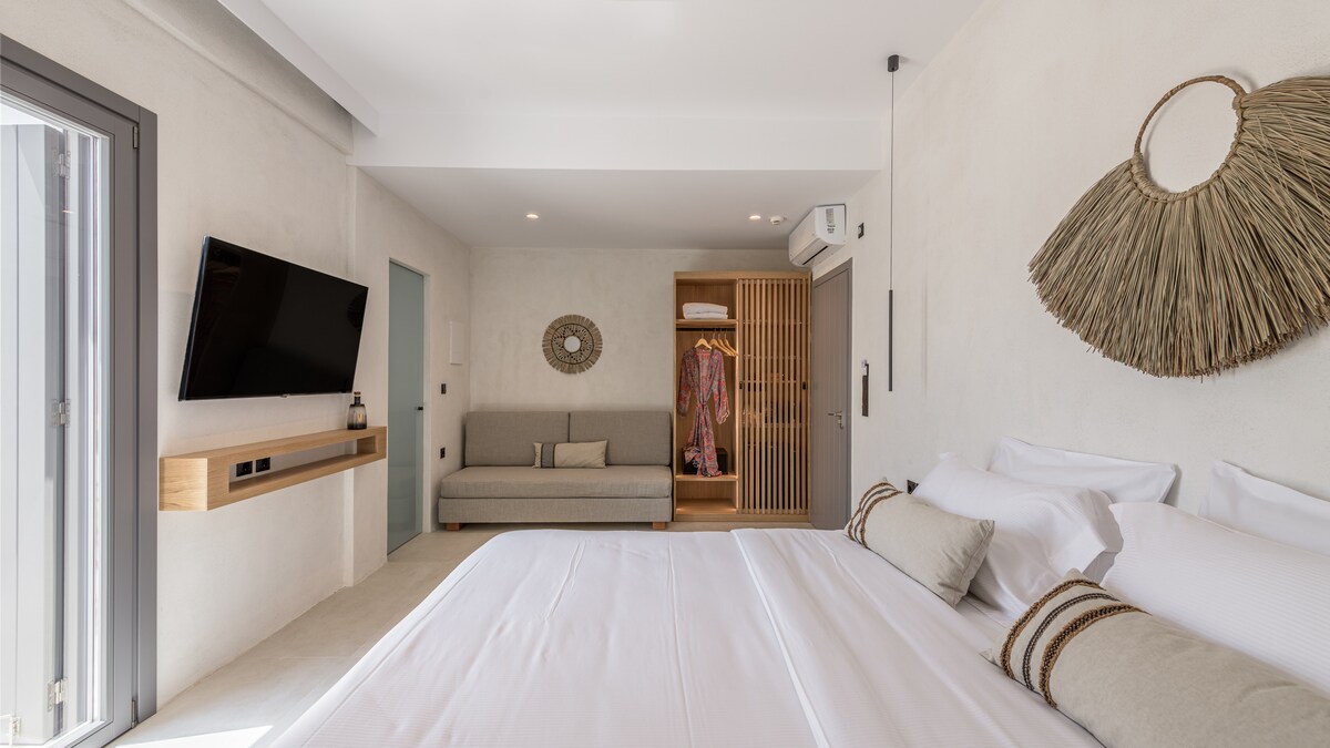 A light-filled bedroom features a spacious bed dressed in white linens with subtle accent pillows. A wall-mounted television is positioned above a wooden shelf, while a cozy seating area is visible nearby. Decorative wall art adds a touch of warmth to the modern space.