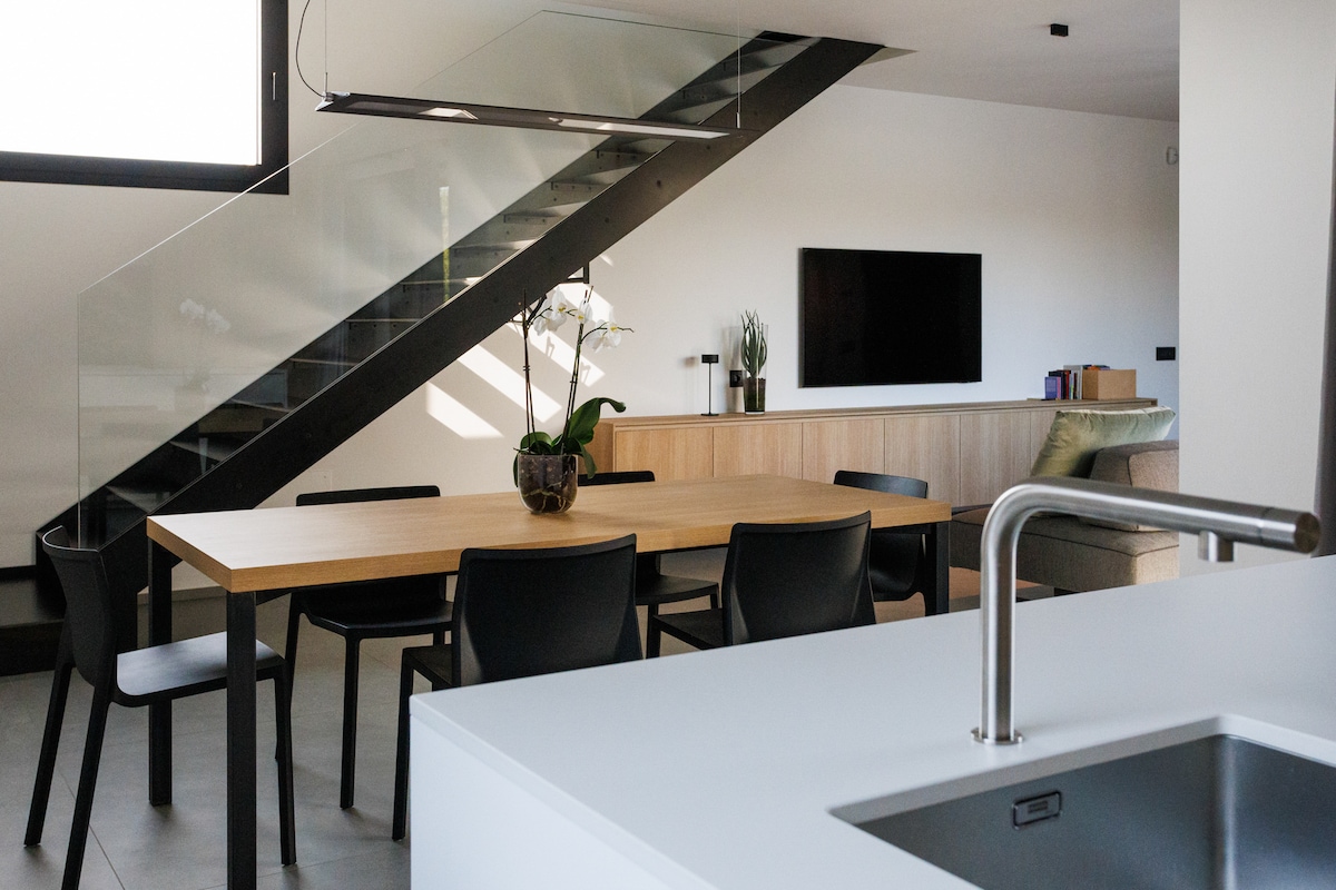 A modern living space features a wooden dining table surrounded by black chairs. A sleek staircase with glass railings leads to the upper level. A minimalistic entertainment unit is positioned against the wall with a television mounted above. Natural light floods the area through large windows.