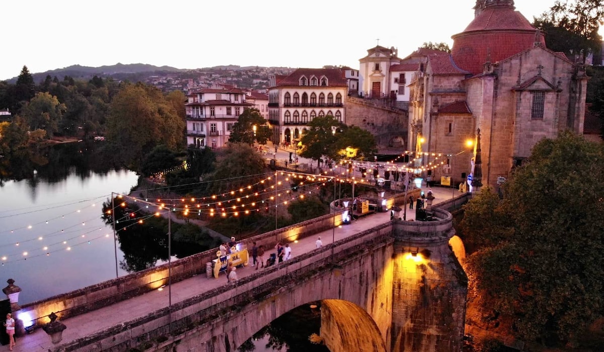 An evening scene features a charming bridge adorned with string lights, illuminating a pathway along the river. Historical buildings are seen in the background, while people gather nearby, creating a lively atmosphere. The setting is enveloped by trees and distant hills, reflecting a serene ambiance.