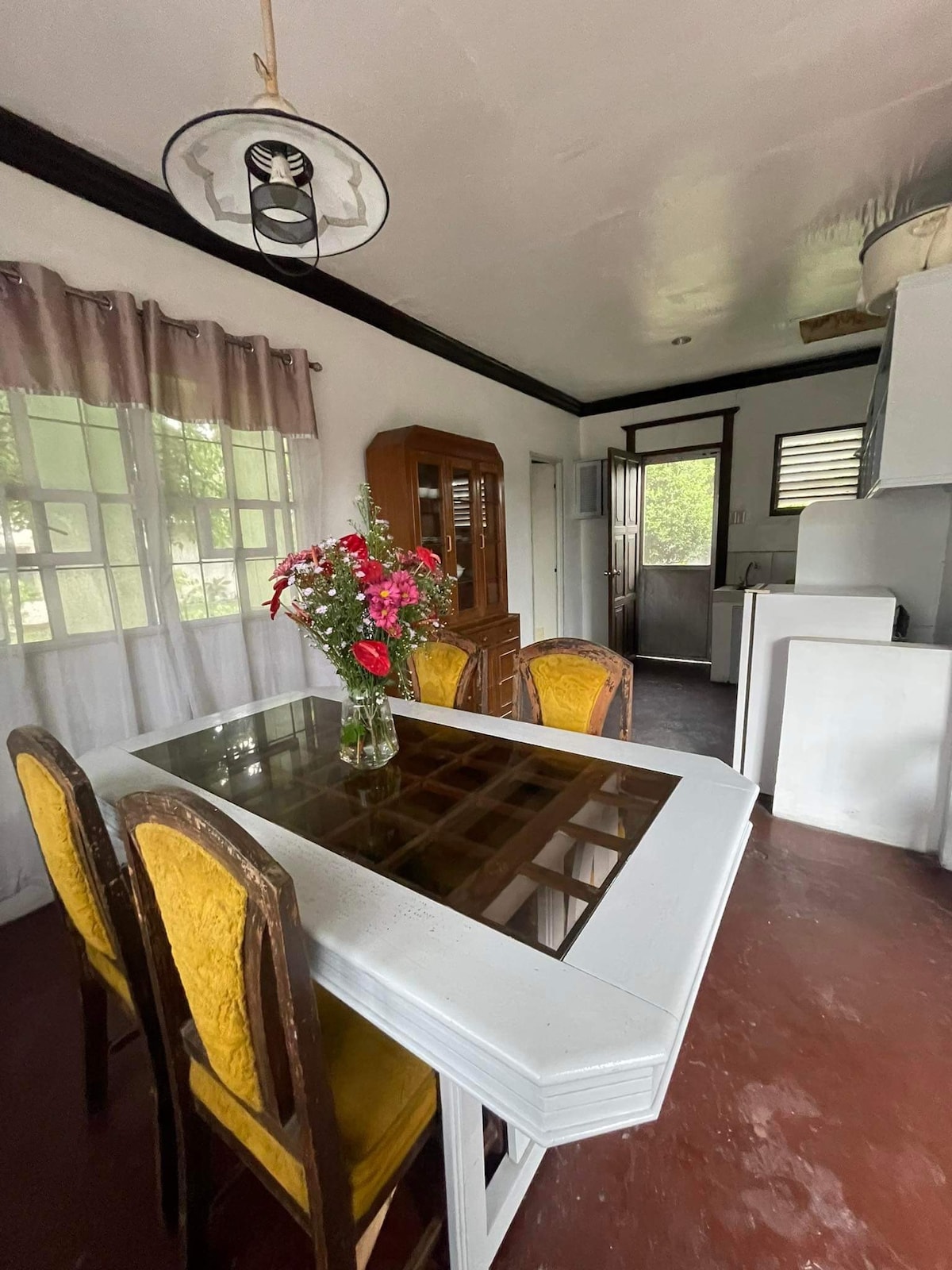 A dining area features a glass-top table surrounded by four yellow-upholstered chairs. A bouquet of red and pink flowers is centered on the table, while natural light filters through sheer curtains. The room includes a wooden cabinet and a door leading outside.