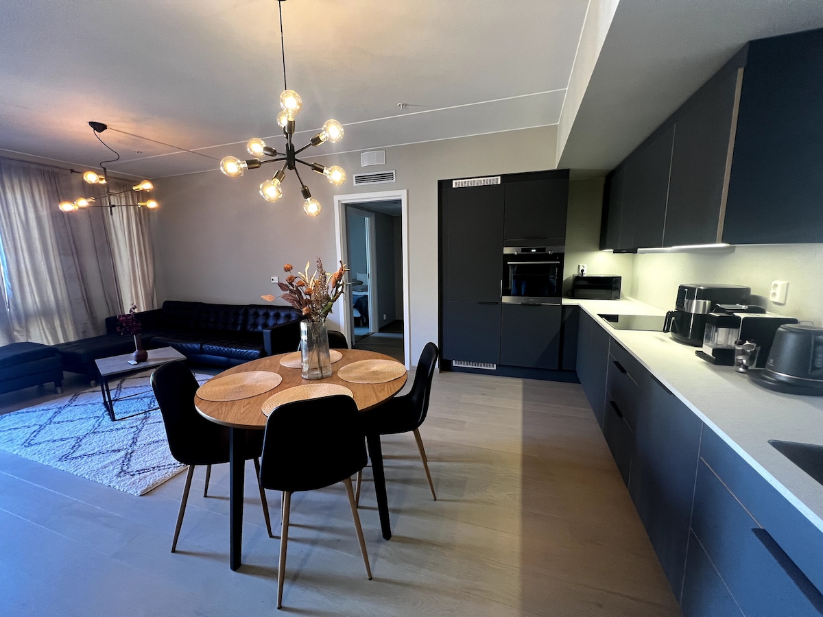 The modern kitchen is equipped with sleek cabinetry and essential appliances, including an oven and coffee machine. A dining table with four black chairs is positioned near a vase of flowers. A cozy living area with a black couch complements the open layout.