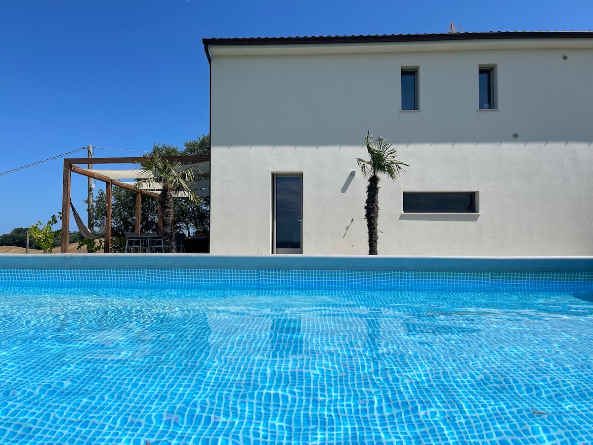 The exterior of a modern building is viewed from a swimming pool, featuring light-colored walls and muted windows. A shaded outdoor seating area is visible, surrounded by greenery and palm trees, with a clear blue sky above.