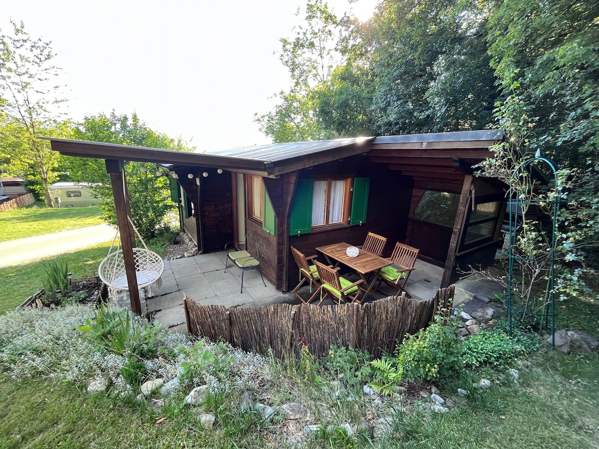 The exterior of the bungalow features a wooden structure with a sloped roof, surrounded by greenery. A patio area includes a table and chairs, alongside hanging lights and a garden with low vegetation. A hammock is seen for relaxation.