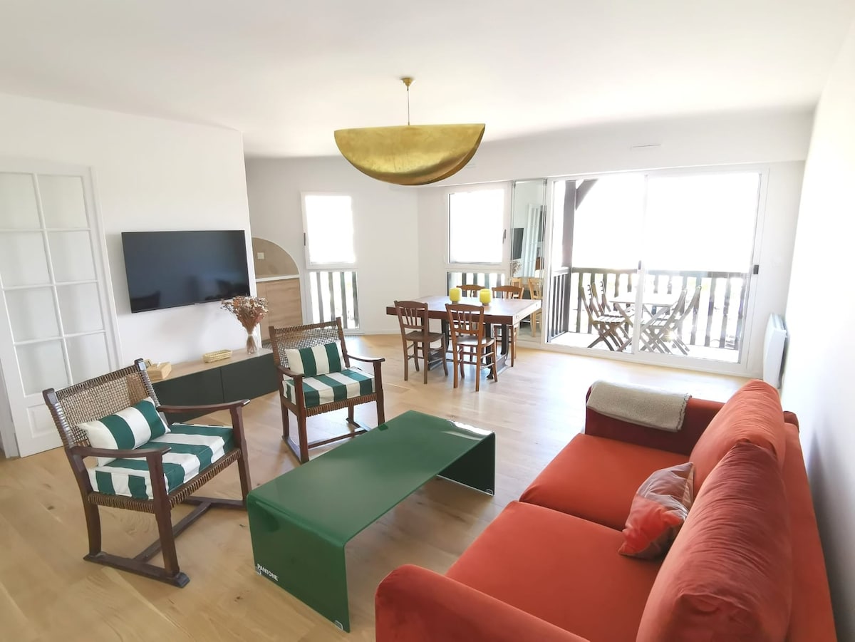 A spacious living area is featured, furnished with a red sofa, striped chairs, and a green coffee table. Large glass doors provide natural light and offer access to a terrace. A dining table is positioned nearby, set against a light wooden floor.