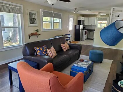 A cozy living area features a dark leather sofa adorned with patterned cushions. A vibrant orange armchair is positioned nearby, along with a blue ottoman. Functional wooden furniture and large windows provide natural light, enhancing the inviting atmosphere. The open layout connects to a well-equipped kitchen.