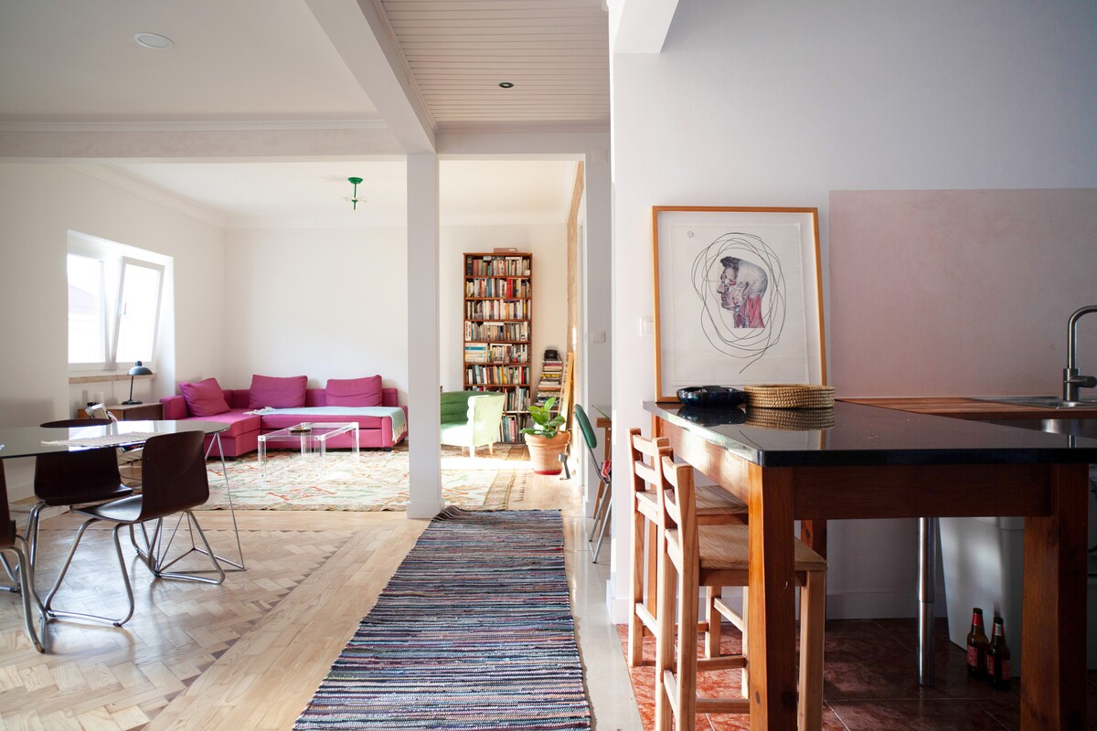 An open living area features a modern design with a pink sofa, hardwood flooring, and large windows allowing natural light. A cozy dining area is visible, equipped with wooden chairs around a table. Artwork adorns the walls, adding character to the space.
