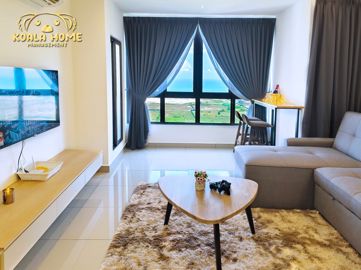 A modern living area features a grey sofa positioned next to a low table on a plush rug. Large glass windows bring in natural light and offer views of the landscape beyond. Dark curtains frame the windows, enhancing the calm environment.
