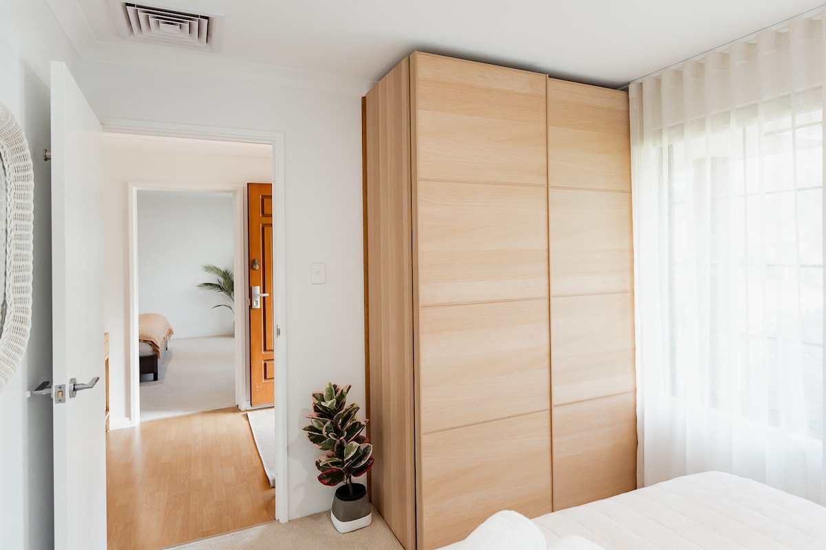 这间明亮的卧室设有带滑动门的大型内置衣柜、舒适的床、透过薄纱窗帘照射进来的柔和自然光线，以及盆栽等装饰品，营造出舒适现代的氛围。