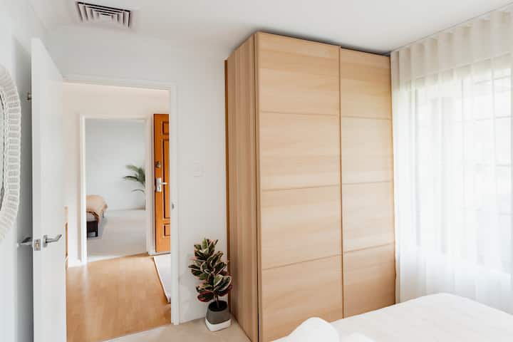 Оваа светла спална соба има голема вградена гардероба со лизгачки врати, удобен кревет, мека природна светлина низ чисти завеси и декоративни детали како саксиско растение за удобно и модерно чувство.