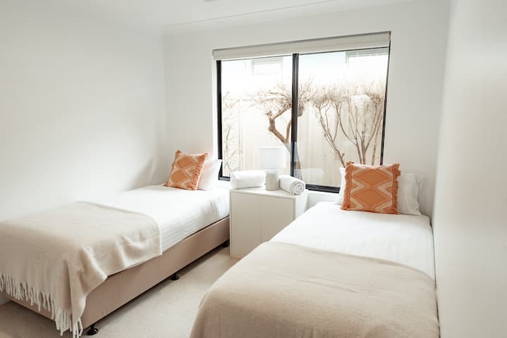 Оваа светла спална соба има два удобни единечни кревета со мека постелнина и декоративни перничиња, заедничка ноќна масичка со чисти крпи и голем прозорец кој нуди природна светлина и поглед на приватен двор.