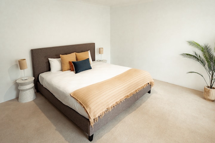 Оваа елегантна спална соба има кадифен брачен кревет со стилска постелнина, ноќни масички со светилки и неутрална палета на бои. Растението во саксија додава малку зеленило, создавајќи мирен и релаксирачки амбиент.