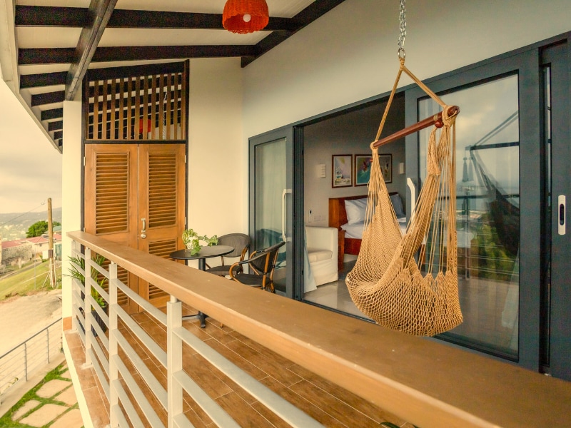A private balcony is featured, showcasing a comfortable hammock and a small seating area. Large sliding glass doors provide access to the room, while wooden accents and a modern railing create a serene outdoor space with partial views of the surrounding landscape.