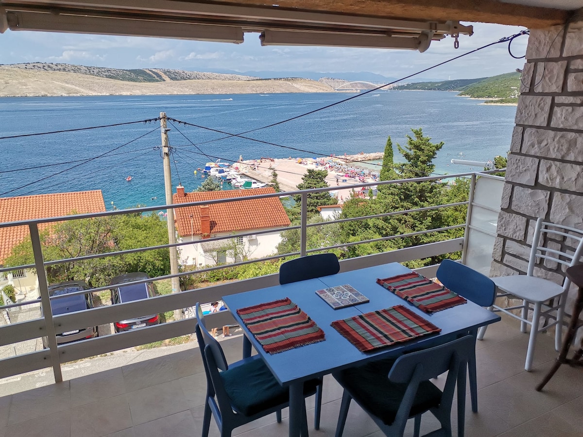 A dining area is situated on a terrace overlooking the sea, featuring a blue table set with striped placemats. Scenic views of the coastline and beach can be seen in the background, framed by greenery and terracotta-roofed houses.