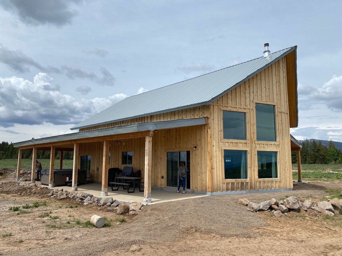 The recently constructed cabin features a wooden exterior with large windows, allowing for ample natural light. A spacious wrap-around porch is visible, providing seating options and access to outdoor views. Scenic surroundings enhance the inviting atmosphere of the cabin.