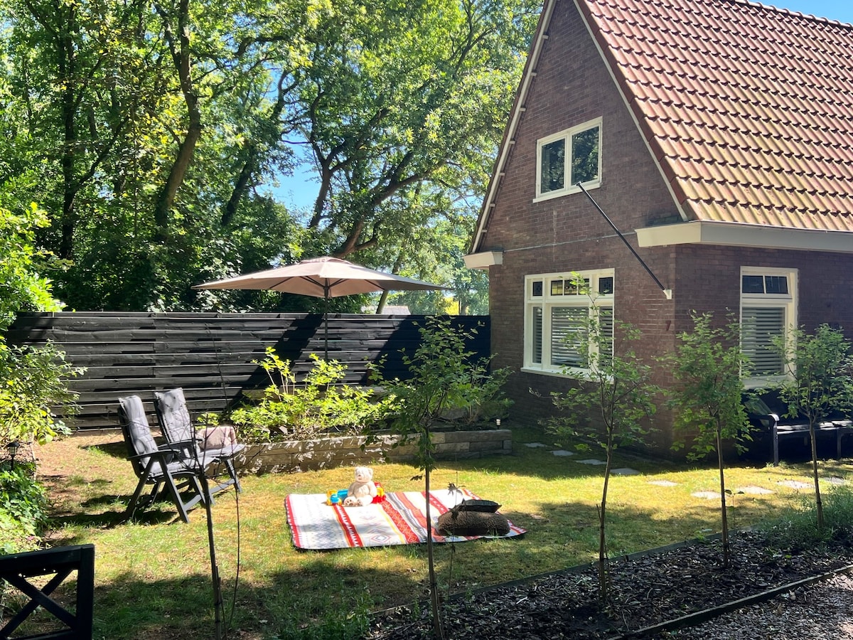 An outdoor area with a well-maintained garden and a cozy blanket on the grass. Two folding chairs are positioned for relaxation under a large umbrella. A brick house with a tiled roof is visible against a backdrop of lush greenery.
