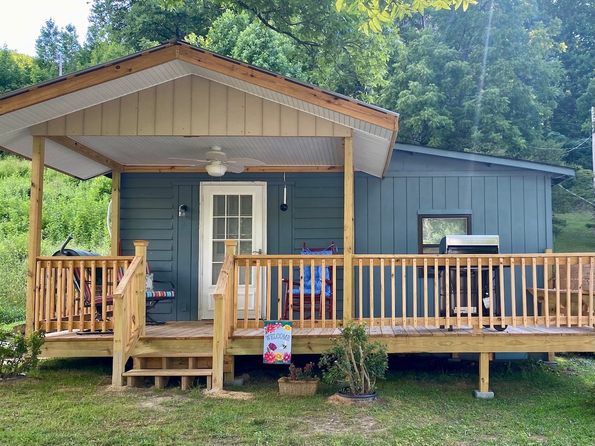 The inviting front porch features a wooden railing and is adorned with a colorful welcome sign. A ceiling fan provides airflow, while a grill and seating area are visible, offering a space for outdoor relaxation surrounded by greenery.