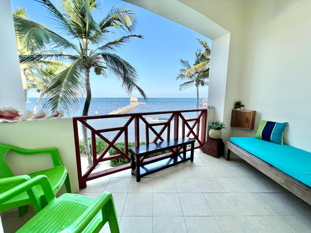 A balcony area overlooks the sea, featuring two green chairs and a wooden bench. Tropical palm trees frame the view, with a wooden dock extending into the water. Light tile floors complement the serene ambiance, offering an inviting space to relax and enjoy the scenery.