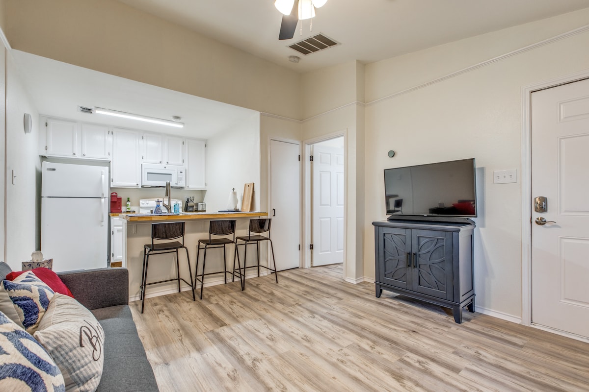 An open living space features a modern kitchen with white cabinetry and appliances. A dining area with bar stools is situated near the kitchen, while a comfortable couch invites relaxation. A flat-screen TV is positioned on a dark wooden cabinet, enhancing the room's functionality.