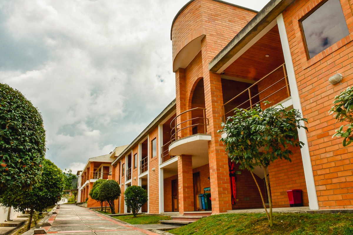 A row of charming brick buildings is seen along a well-maintained pathway. Lush shrubs and small trees are positioned on either side, providing greenery. A covered balcony is featured on the second floor of one building, with a cloudy sky serving as a backdrop.