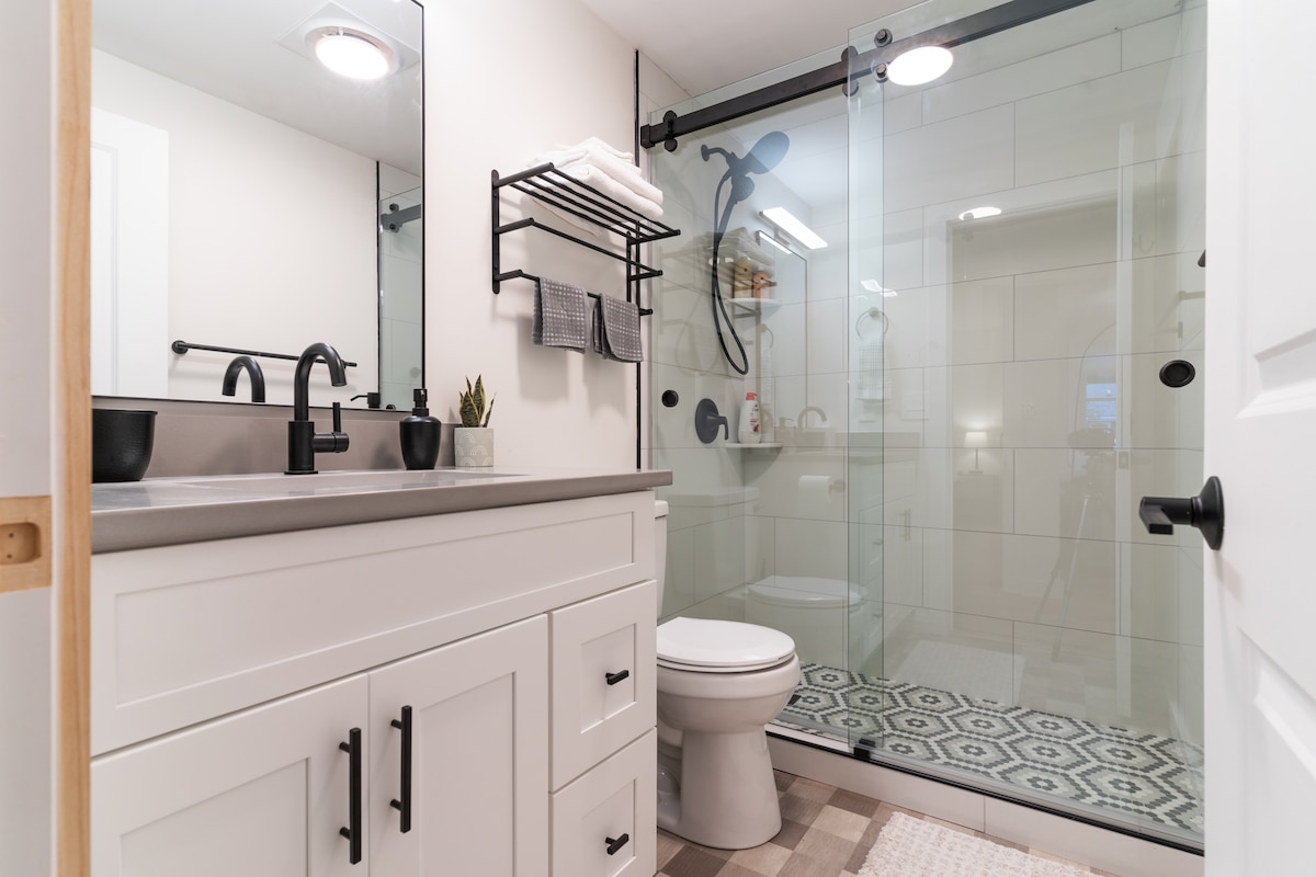 A modern bathroom features a sleek, glass shower enclosure with black fixtures and a patterned floor. A white vanity with black hardware offers storage, and shelves are neatly arranged with towels. A small plant adds a touch of greenery to the space.