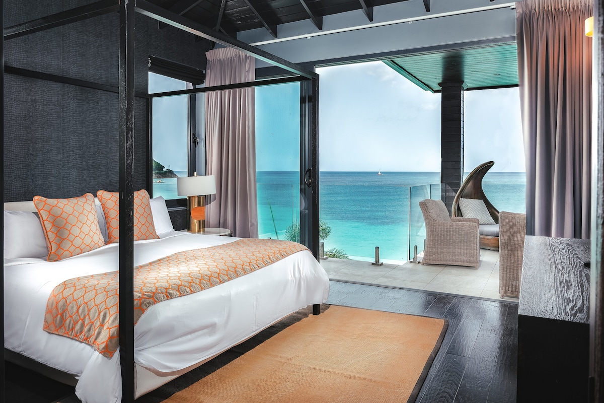 A spacious bedroom features a four-poster bed dressed in white linens with orange accent pillows. Floor-to-ceiling windows provide an ocean view, complemented by a comfortable seating area with a chair and side table. Natural light fills the room, enhancing the sleek design.