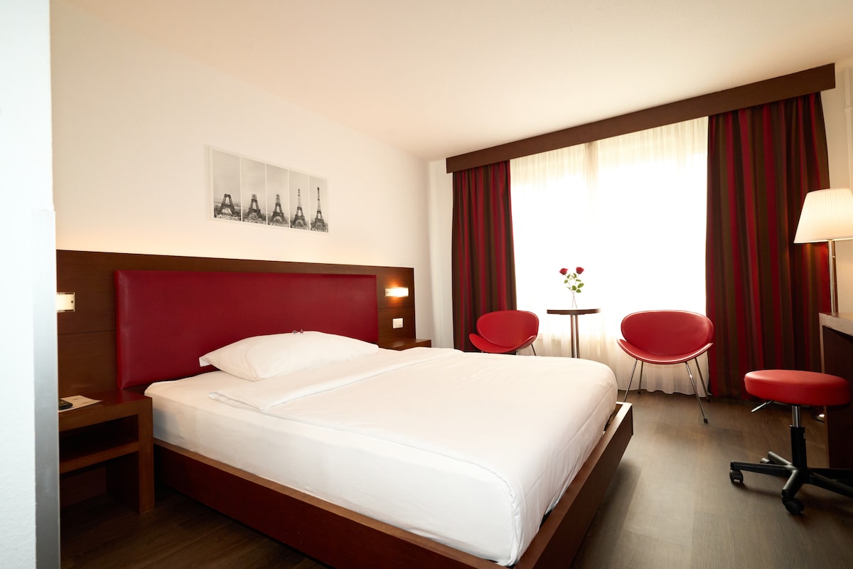 A spacious room features a large bed with white linens and a wooden platform. Two red chairs and a small table provide seating. Natural light filters through large curtains, illuminating the area. A framed photo of the Eiffel Tower adorns the wall.