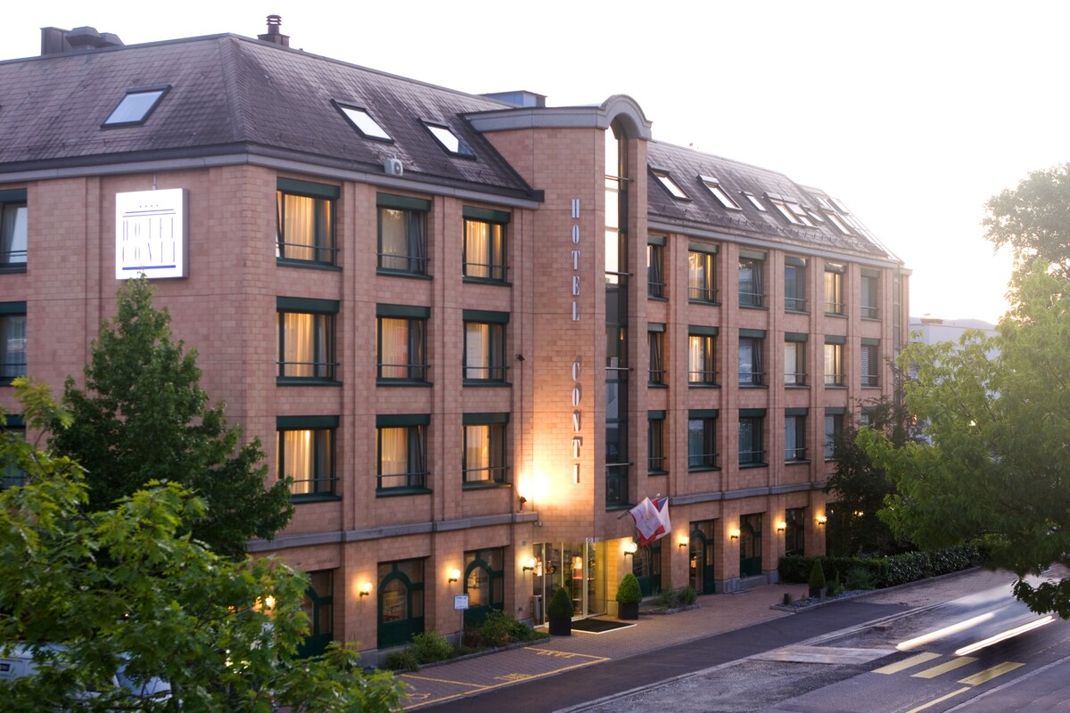 The exterior of the hotel features a brick facade with large windows. The entrance is adorned with flags, and the building is framed by greenery. Soft lighting highlights the hotel signage as it stands against a twilight sky.