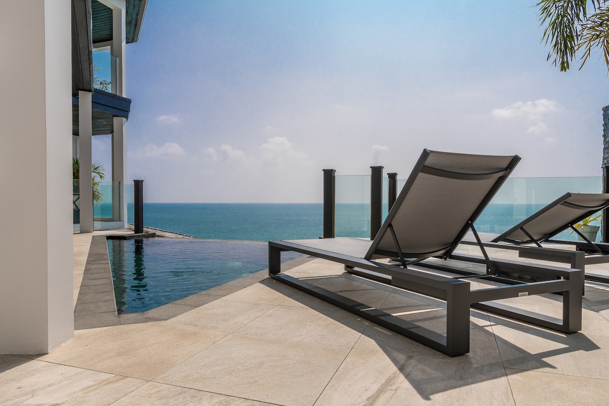 Lounge chairs are positioned beside an infinity edge pool, overlooking the expansive Caribbean Sea. Smooth stone tiles create a seamless transition to the water’s edge, while the horizon is bathed in natural light, enhancing the serene environment.