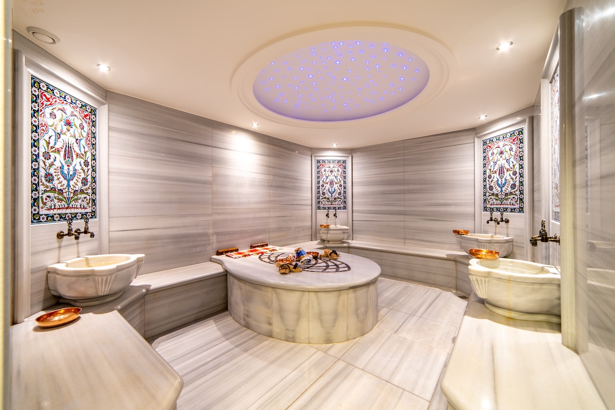 A serene spa-like environment is presented, featuring smooth marble surfaces and intricate decorative tiles on the walls. Ambient lighting is provided by a starry ceiling feature, complementing the soothing arrangement of wash basins and seating areas.