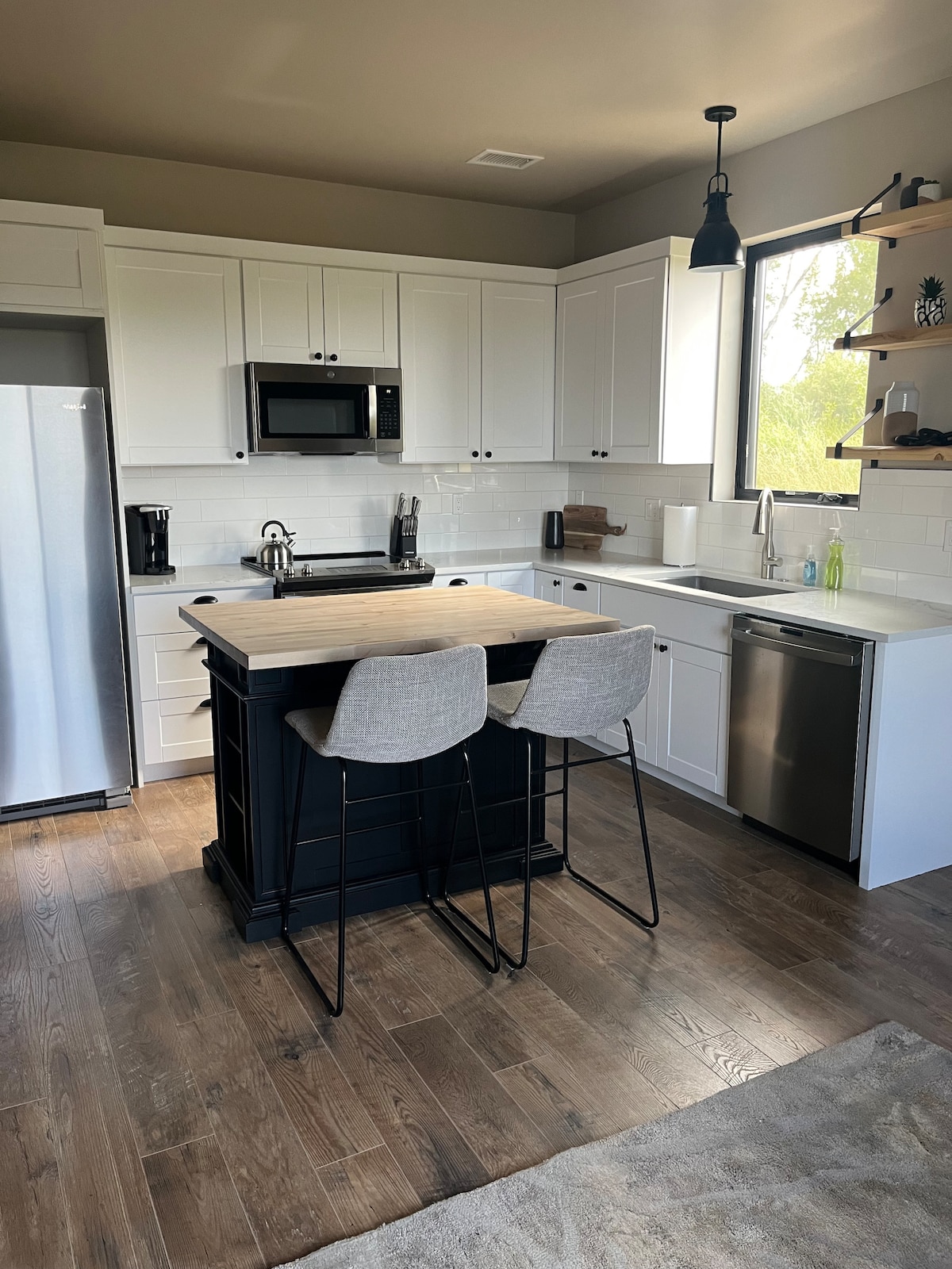 A modern kitchen is equipped with white cabinetry and stainless steel appliances, including a refrigerator and microwave. A small island with two gray bar stools provides additional seating. Natural light enters through a window, illuminating the workspace and a pleasing arrangement of kitchen essentials.