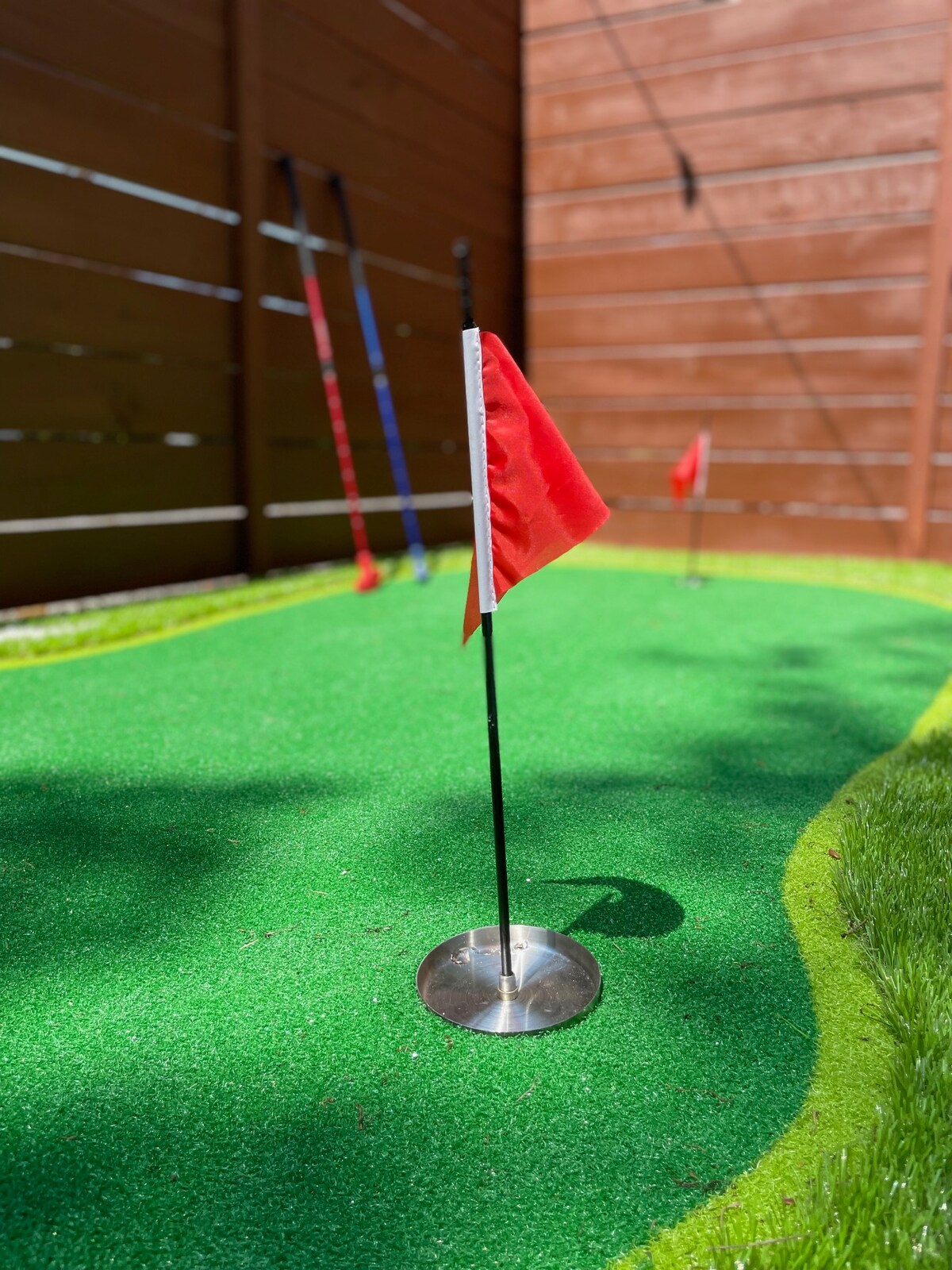 A putting green is depicted, featuring several red flags positioned in silver bases. The vibrant green turf provides a smooth surface for practice, surrounded by a neatly trimmed edge. The wooden fence in the background adds a sense of enclosure to the area.