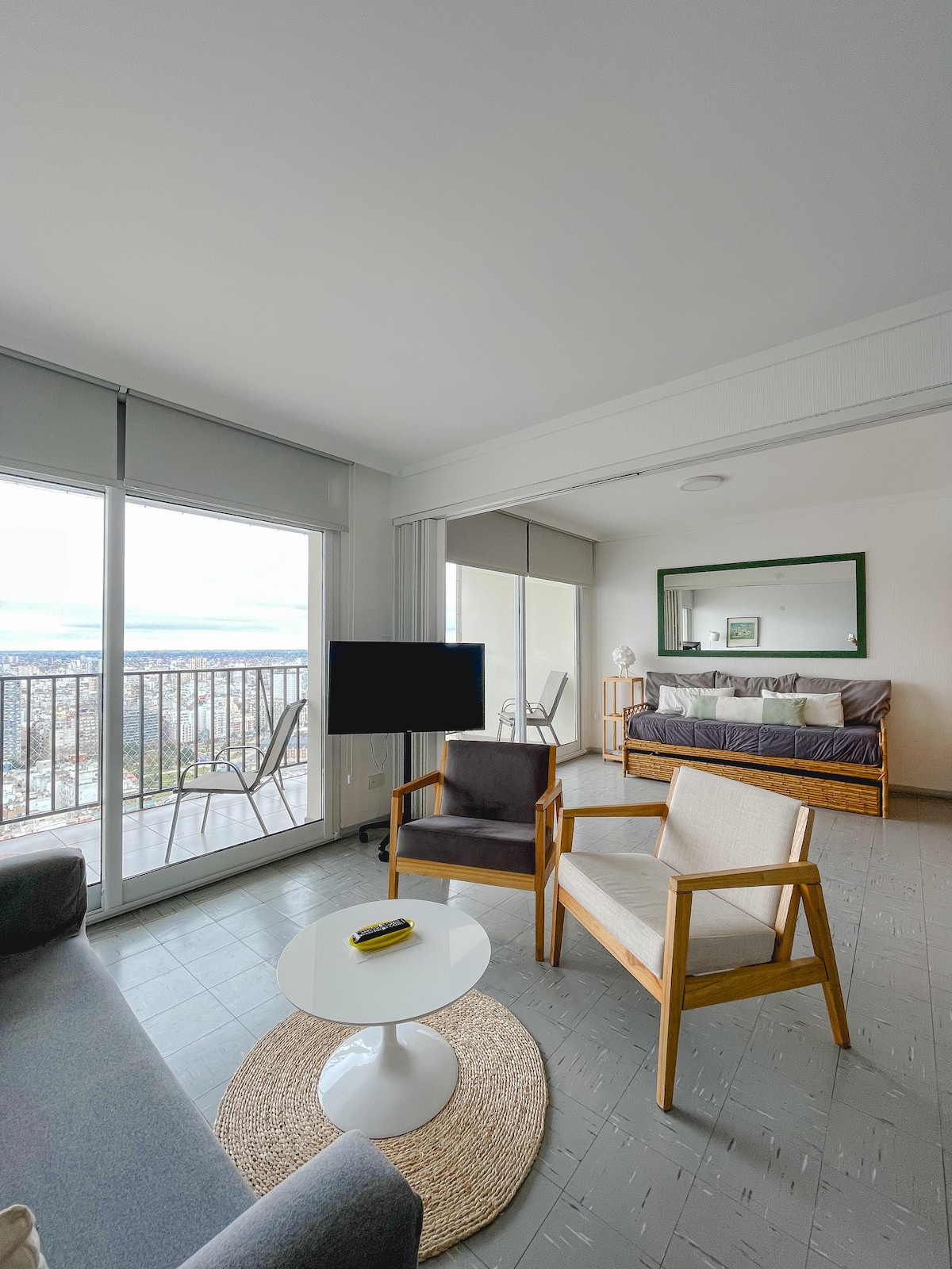 A bright living area is displayed with large windows offering expansive views. Two armchairs and a coffee table are arranged around a light rug. In the background, a spacious bed is framed by sliding glass doors leading to a balcony.