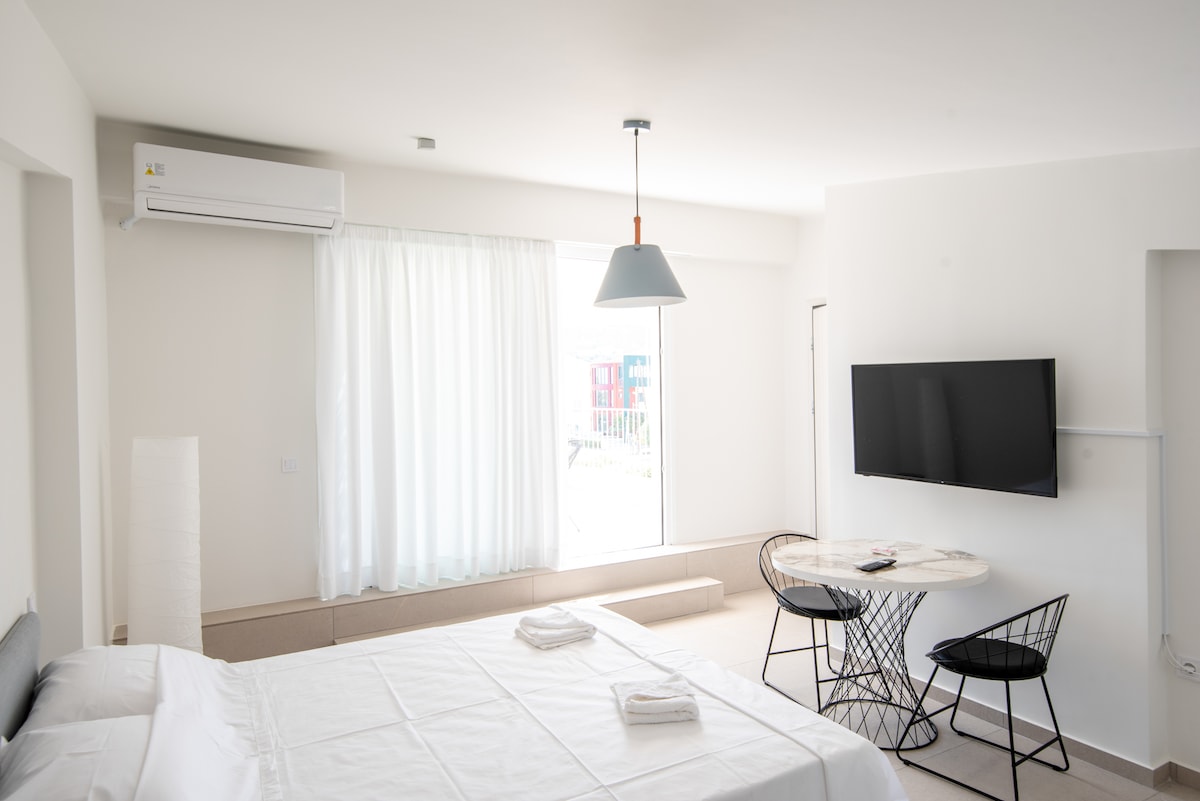A light-filled room features a spacious bed with neatly arranged linens. A round table set for two is paired with modern wire chairs. Large windows with sheer white curtains allow natural light to illuminate the neutral tones of the space.