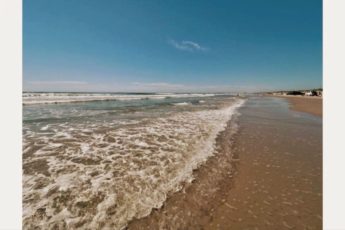 A serene beach landscape is depicted with gentle waves lapping against a sandy shore, under a clear blue sky. The horizon showcases distant beachgoers enjoying the sun, while the foamy water creates a tranquil atmosphere.