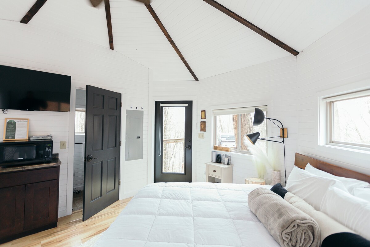 An octagon-shaped room features vaulted white ceilings with exposed wooden beams. A spacious bed is complemented by a soft blanket. A small kitchenette with a microwave is visible. Natural light enters through multiple windows, and a door leads to a private outdoor area.